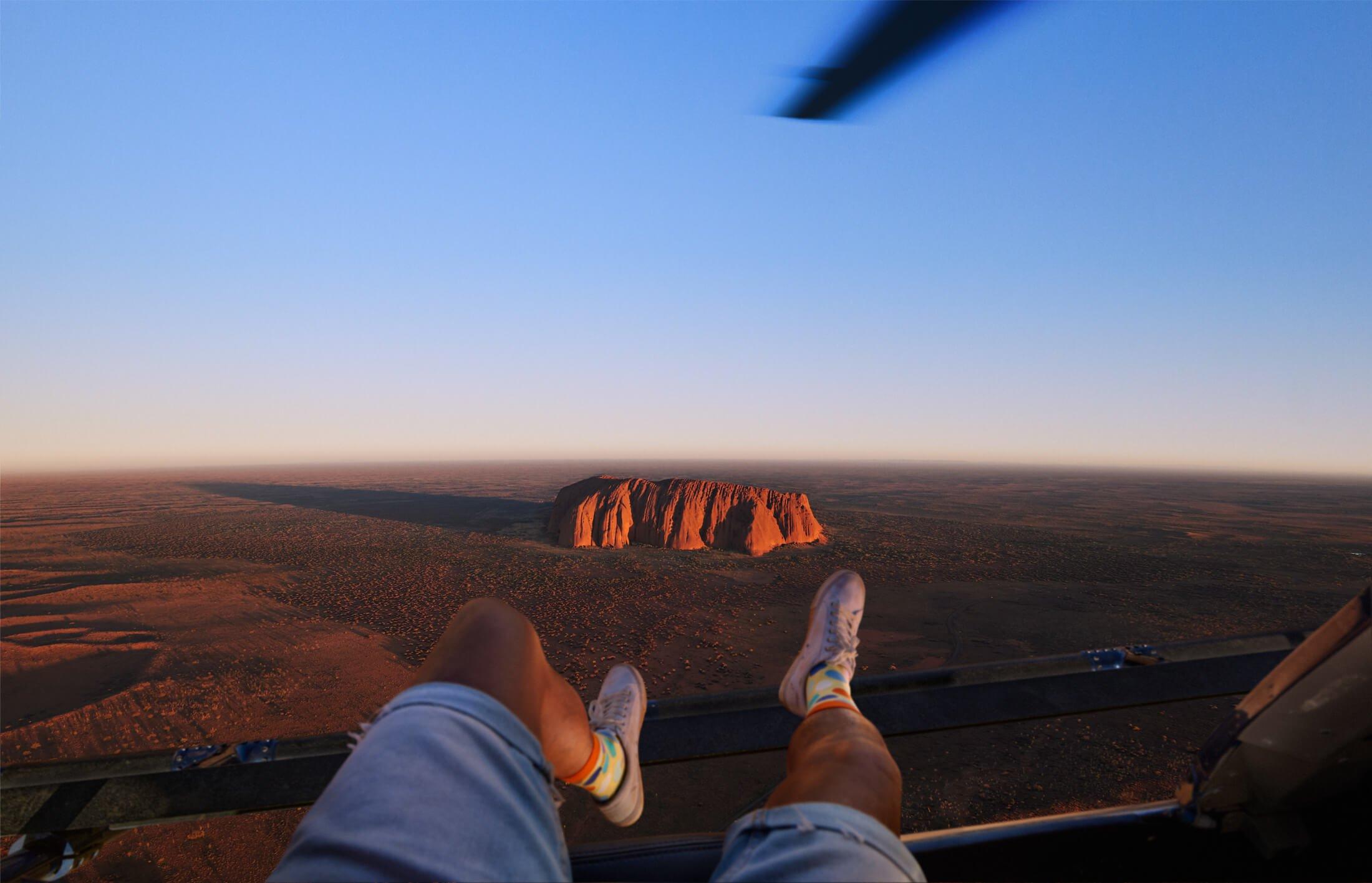 Point Of View of a person sitting on a helicopter dangling their legs over the edge with Uluru down below