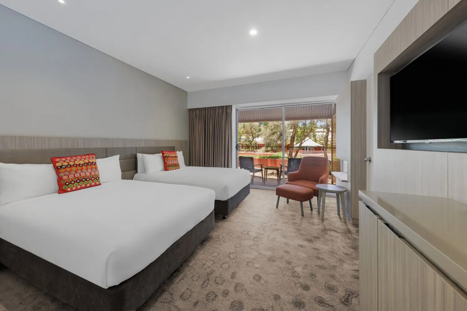 Two double beds with white sheets, TV mounted to the wall looking out towards a balcony with outdoor chairs
