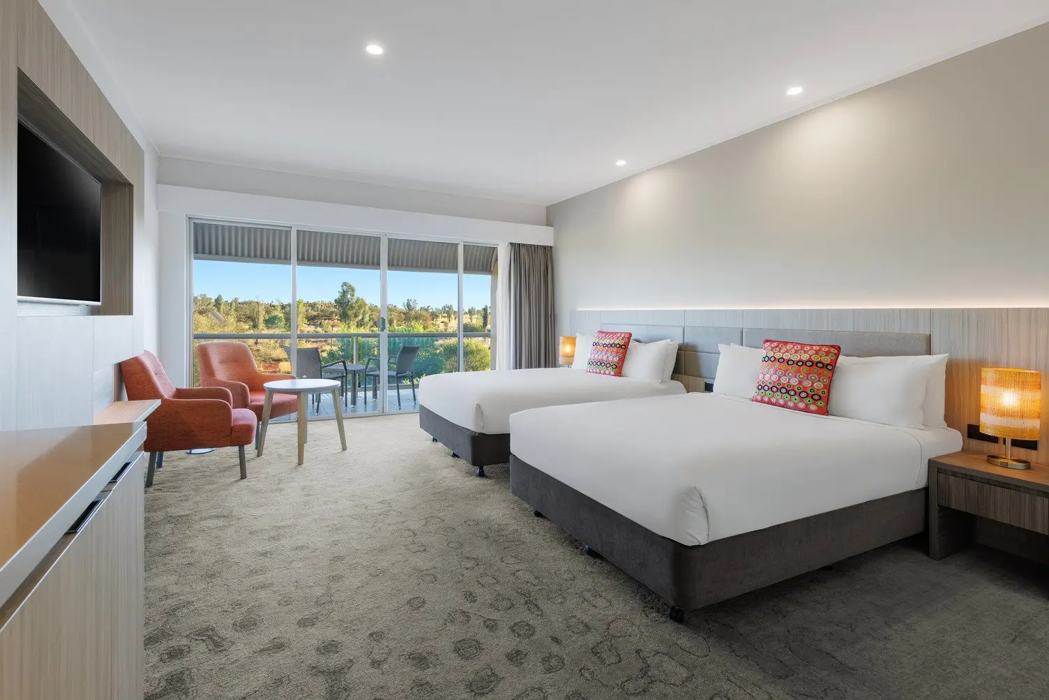 Two double beds with a TV mounted to the wall, two comfortable chairs and an outdoor area looking onto the desert lanscape