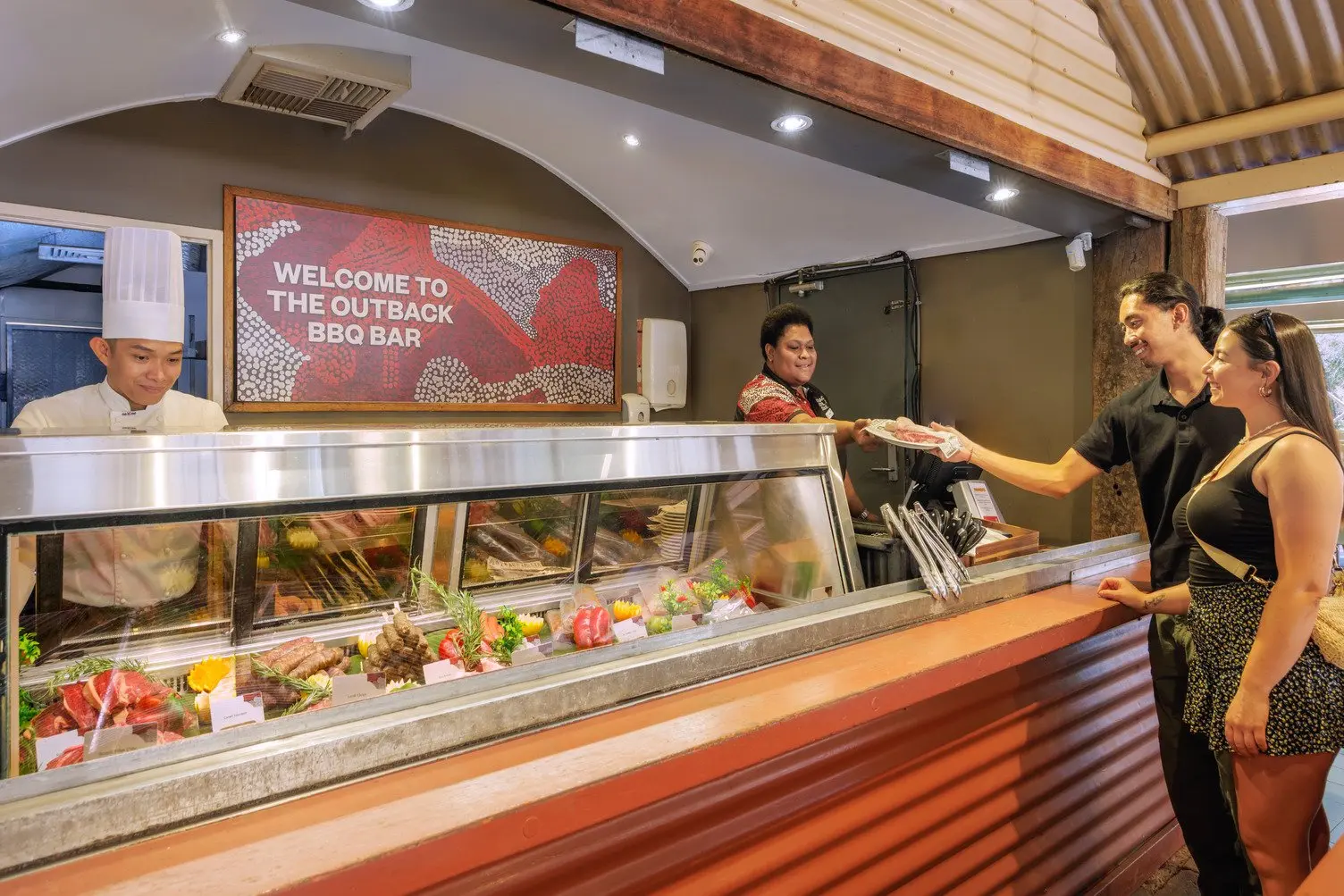 Two women getting handed a piece of meat by staff in front of the meat selection and chef