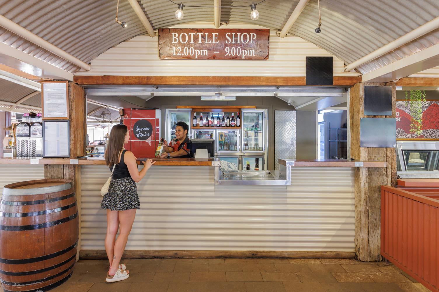 A women who works out the bottle shop is showing a customer a bottle of wine