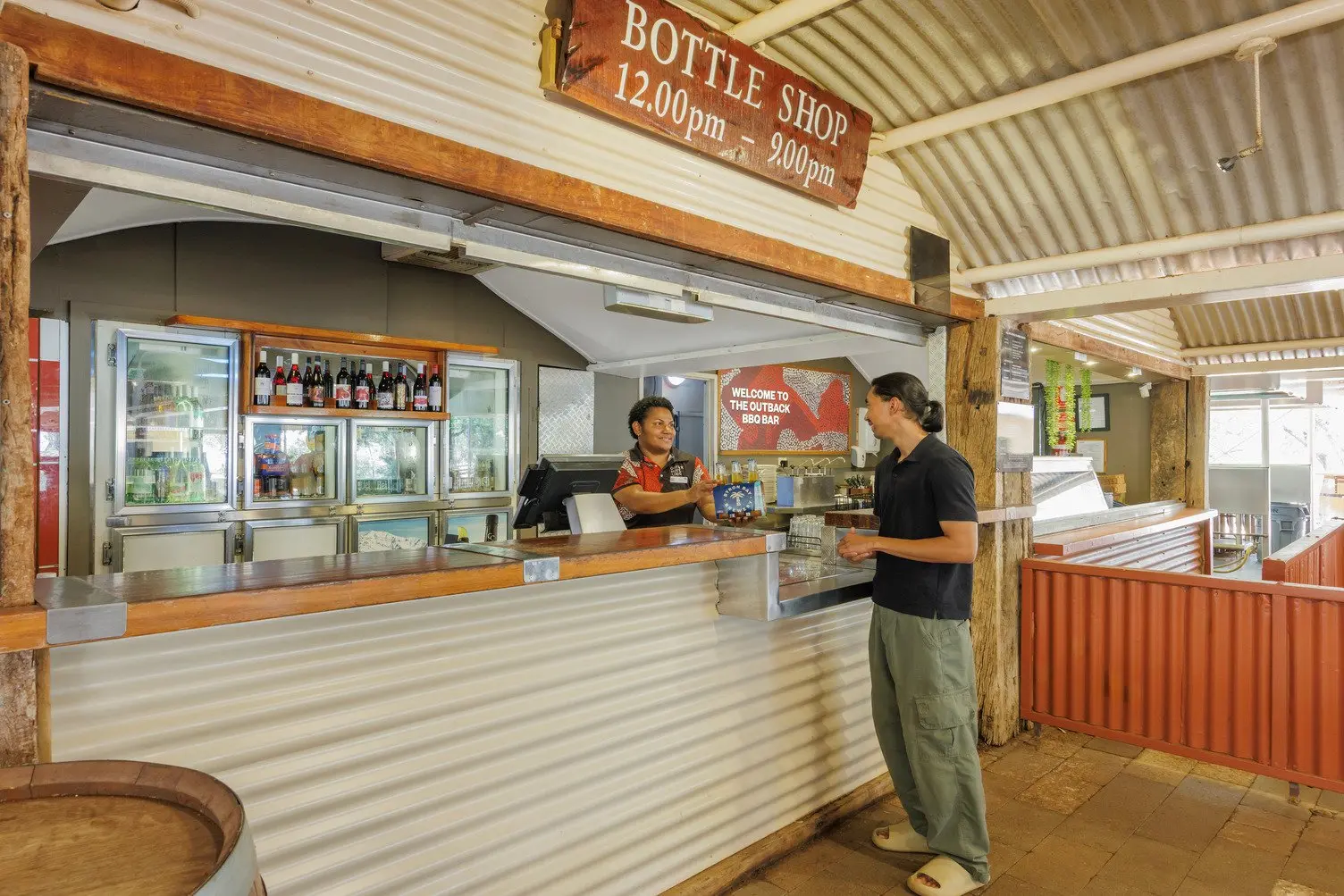A women is passing a 6 pack of Byron Bay Brewery beers to a customer