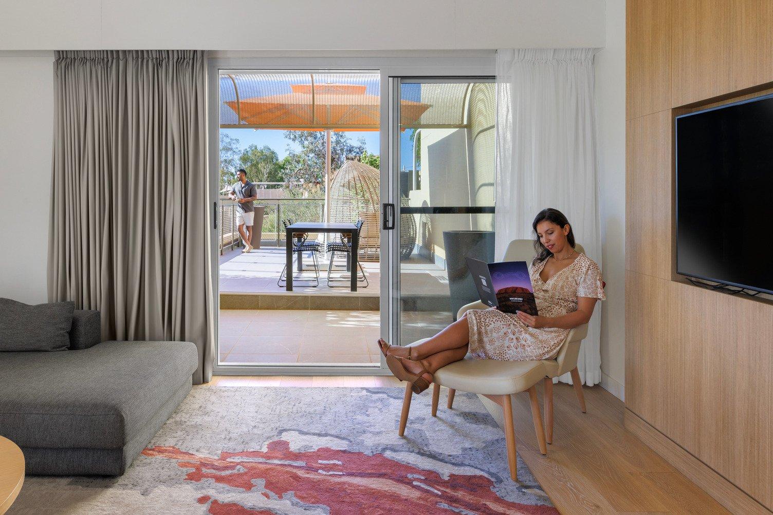 Women read a Ayers Rock Resort Magazine sitting on a chair while a man stands outside on the balcony looking outward