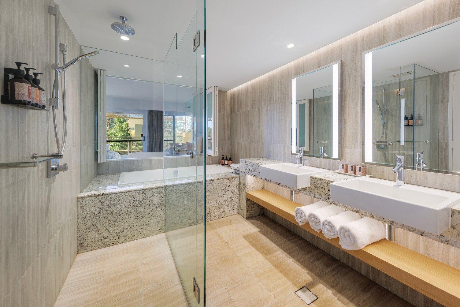 Large bathroom with 2 large mirrors, double sink, shower and bath looking onto the bedroom and balcony
