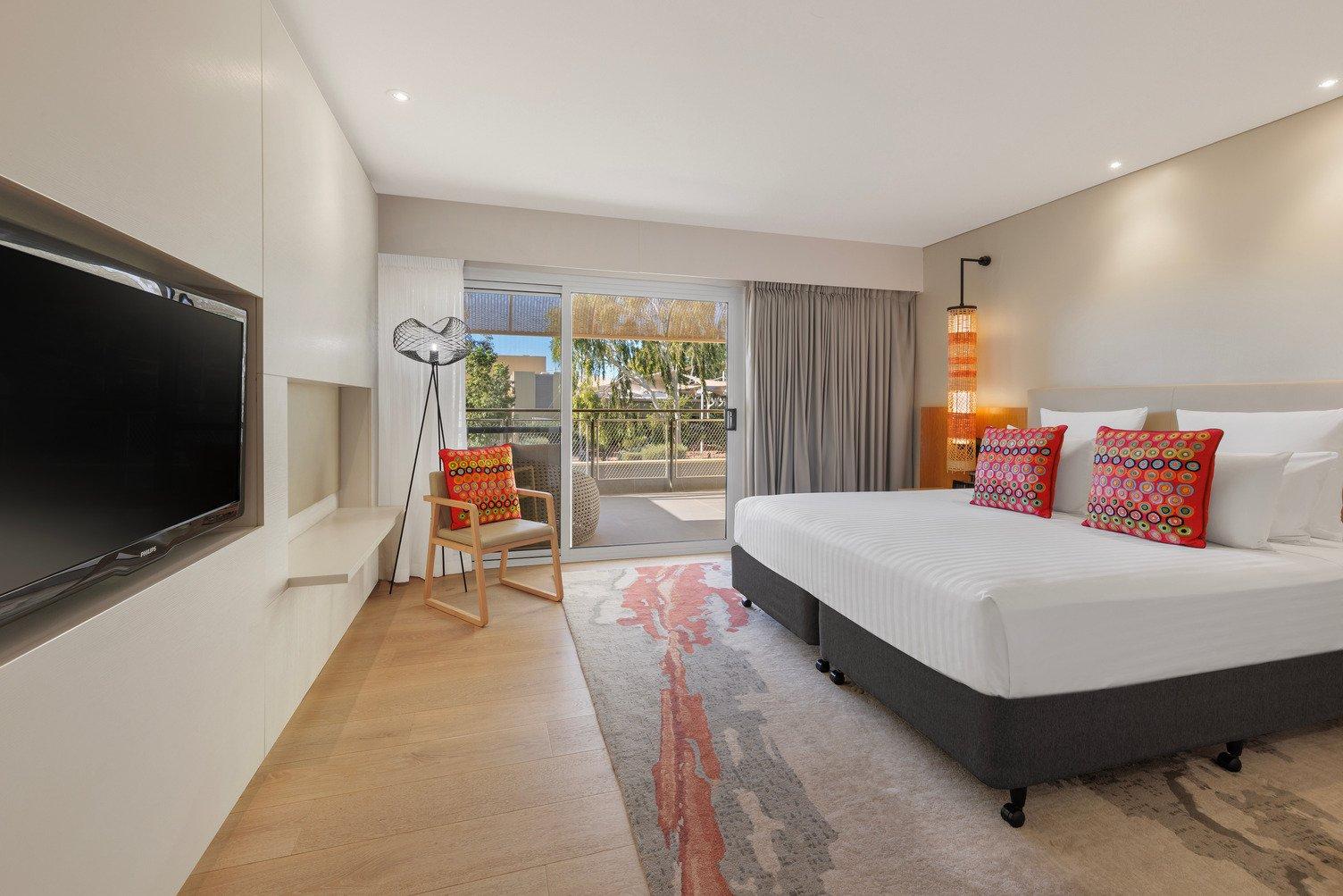 Large bed with white sheets, TV mounted to the wall, looking onto the balcony with open grey curtains