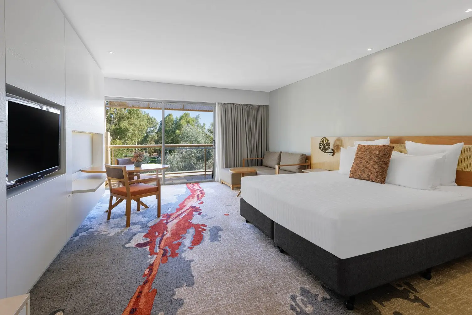 A king bed with white sheets, grey couch near the balcony with curtains open, table and chairs, and TV.