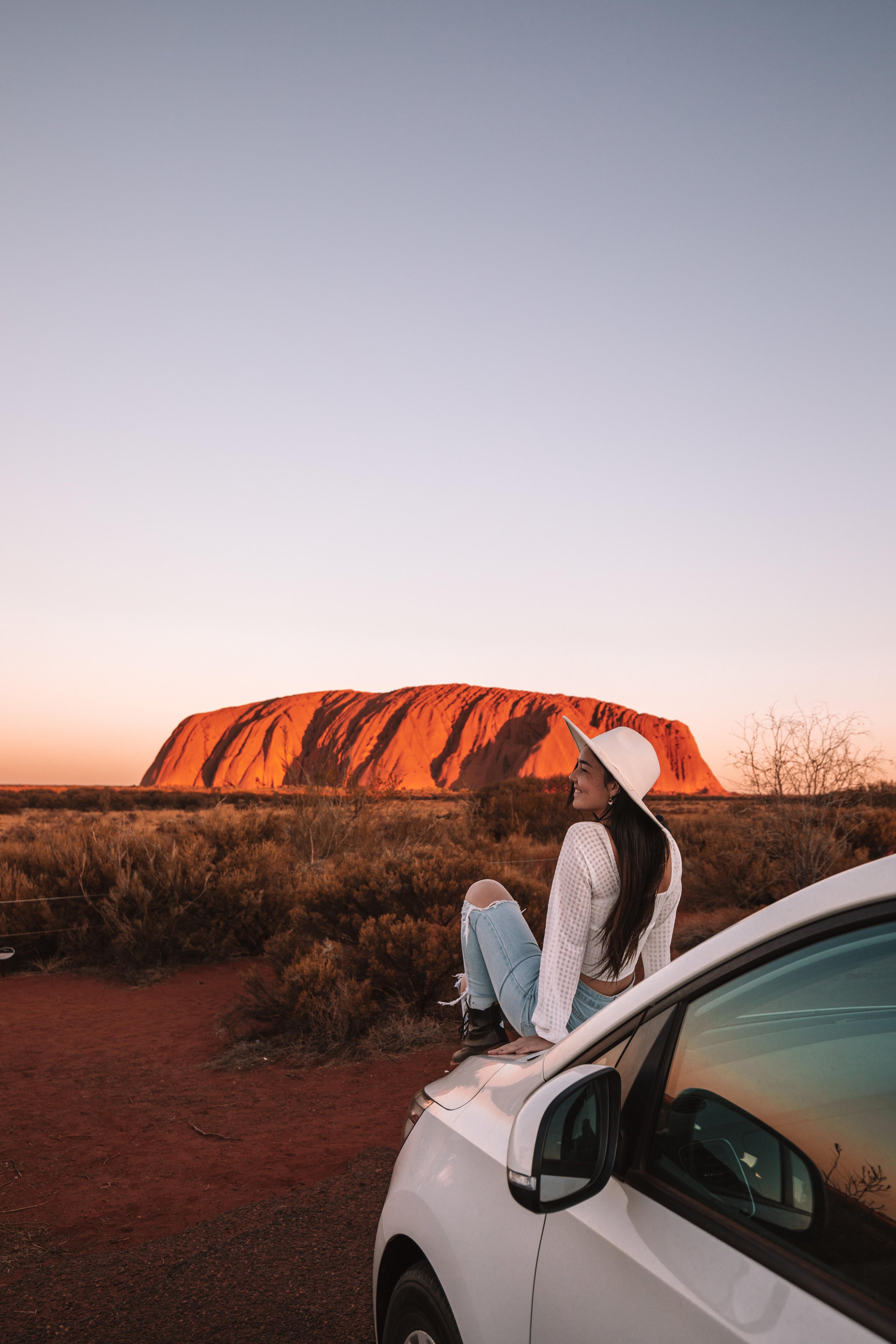 Woman sitting on car hood in front of Ayers Rock