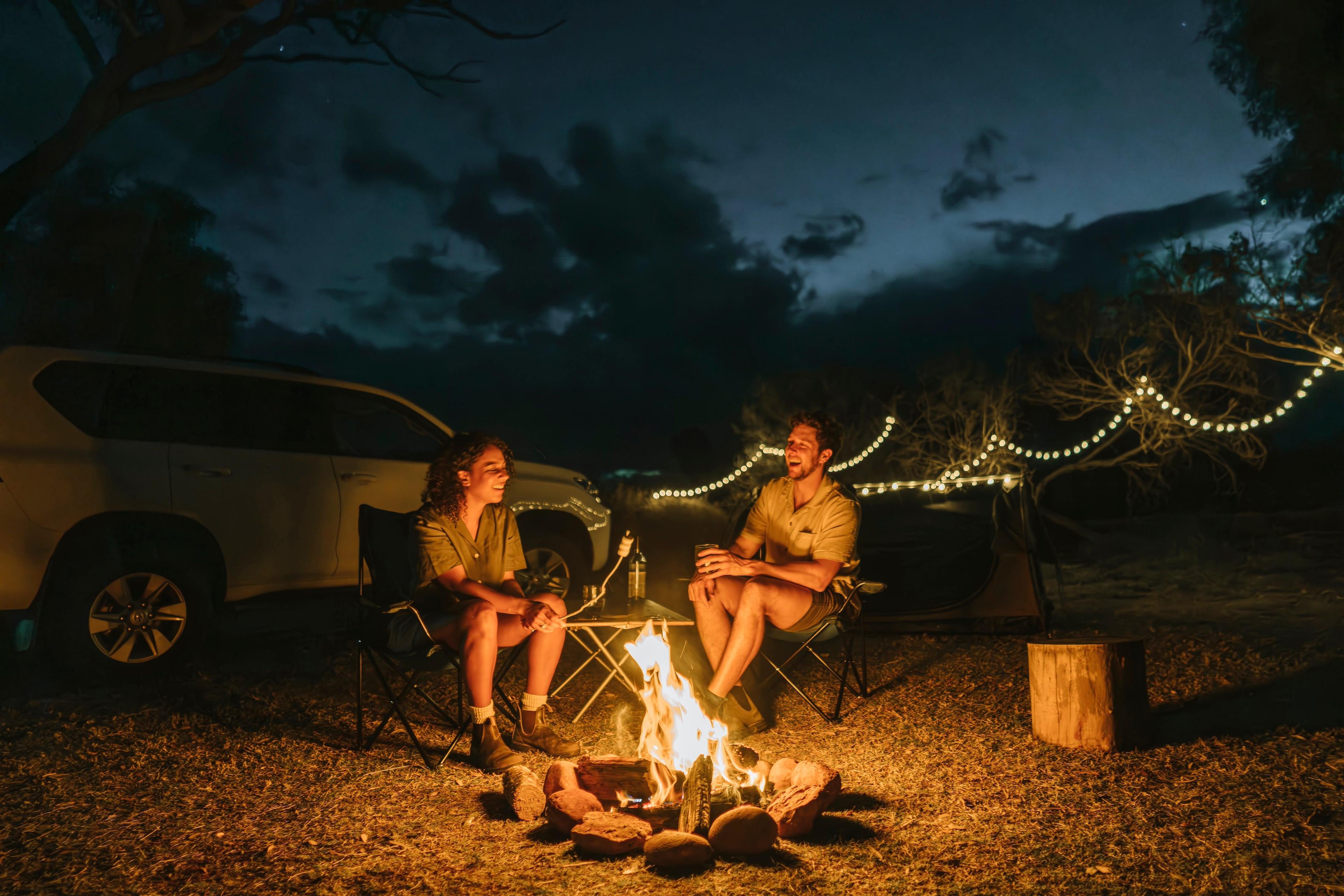 A couple enjoys a campfire at night, at a campsite set up with string lights