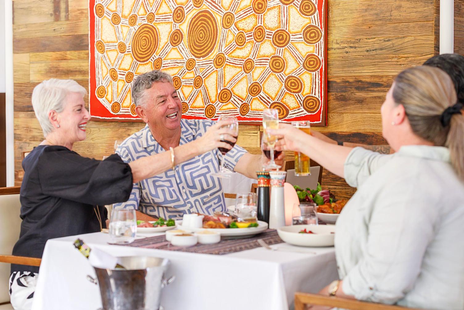 A group of 4 people raising their glasses and smiling before they enjoy their meal at Arnguli Grill. Behind the people details a beautiful Indigenous painting.