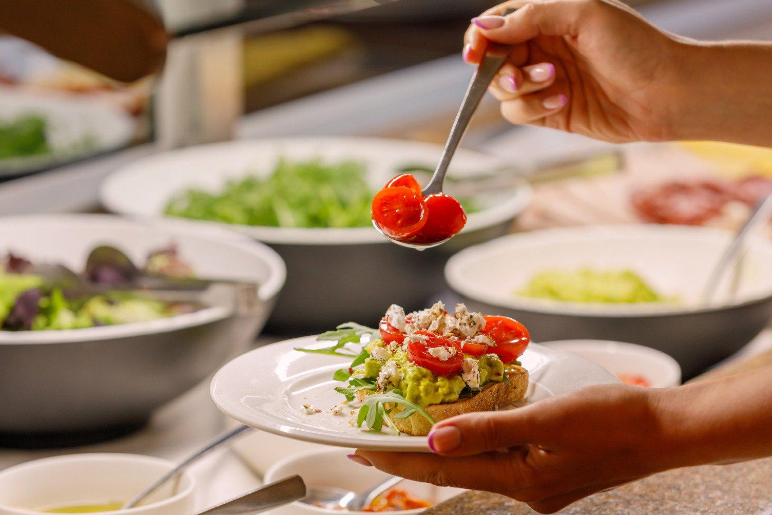 A person holding a plate with avocado and tomato on toast in front of the buffet at Bough House Restaurant