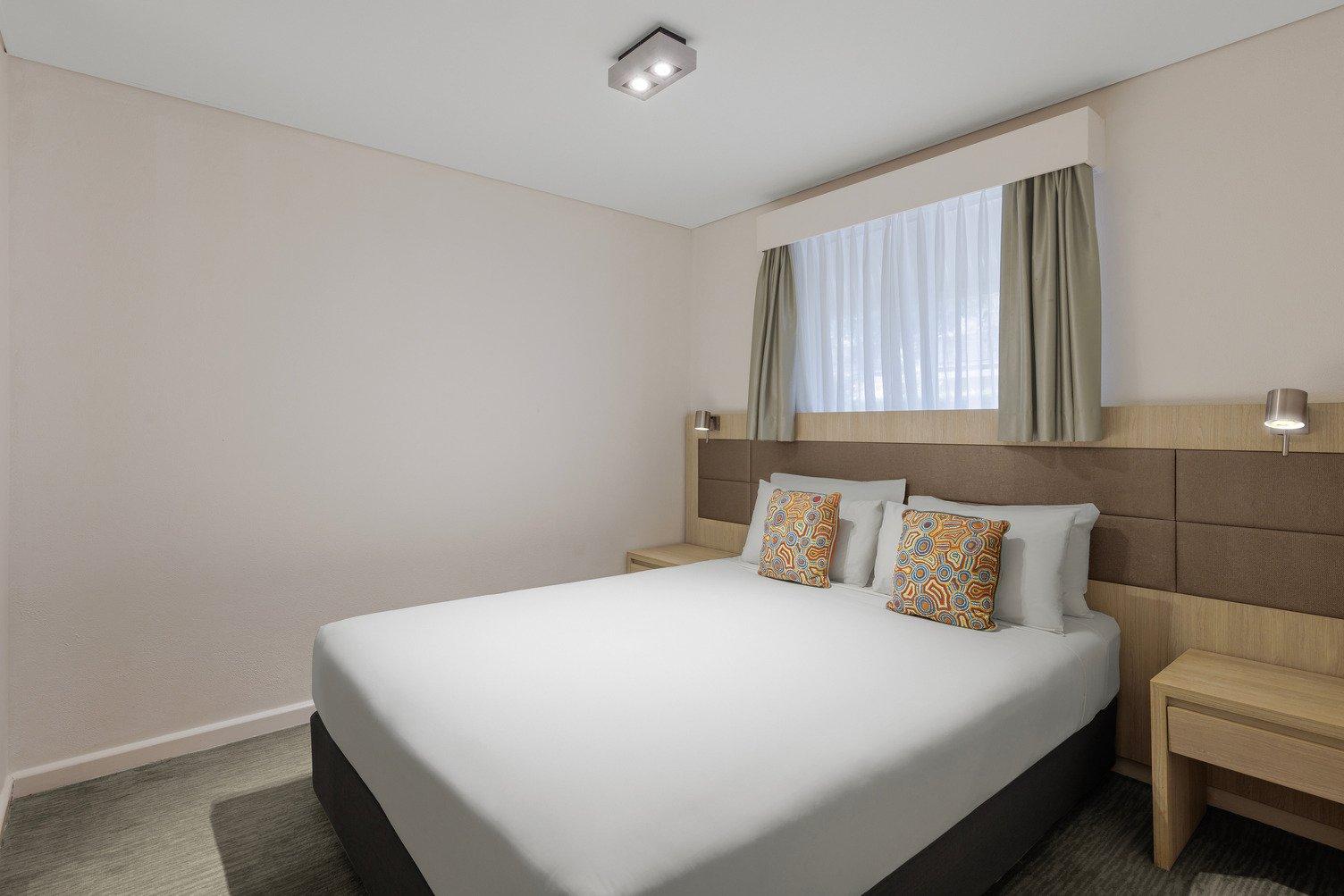 Queen bed with white sheets and two cushions, with a window above the bed frame