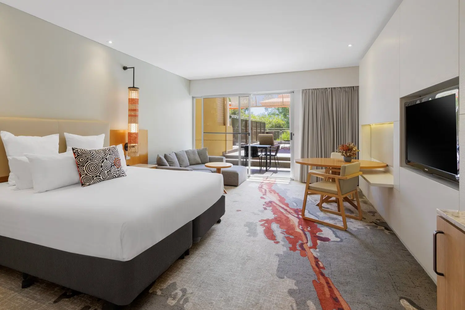 King bed with white sheets, TV mounted to the wall, L shaped grey lounge looking onto balcony with shares