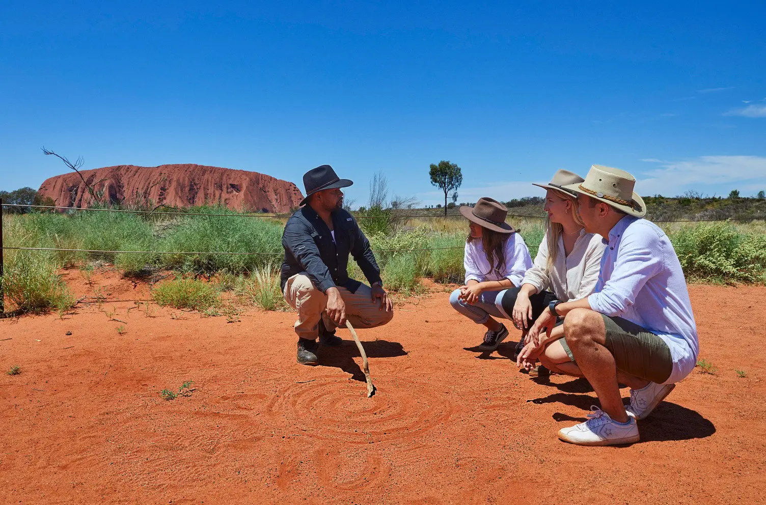 a guide is kneeling and draws a waterhole in the red dirt with Uluru in the background. 3 people look at the guide kneeling.